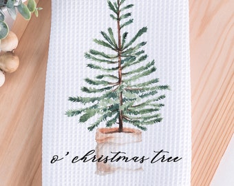 May include: White waffle-weave kitchen towel featuring a watercolor illustration of a Christmas tree in a pot. The tree is green with a brown trunk, and the pot is a light brown color. The text "o' christmas tree" is written in black script.