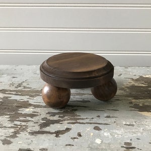 May include: A small, round, wooden plant stand with a dark brown finish. The stand has a circular top supported by three spherical legs. The wood grain is visible, adding to the natural aesthetic. This decorative stand is suitable for displaying small plants or decorative items.