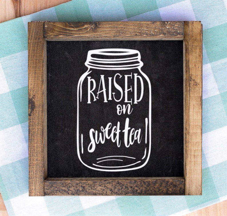May include: A square wooden sign with a dark background and a white mason jar illustration. The words "RAISED on sweet tea" are written in a script font within the jar. The sign is framed with a dark brown wood.