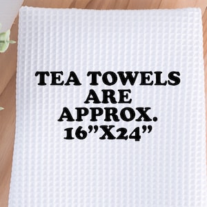 May include: White waffle-weave tea towel with the text "TEA TOWELS ARE APPROX. 16"X24"" in black. The towel is rectangular and has a textured surface. The background is a light wood surface.