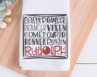 May include: White waffle-weave kitchen towel with a black and red graphic. The graphic lists the names of Santa's reindeer: Dasher, Dancer, Prancer, Vixen, Comet, Cupid, Donner, Blitzen, and Rudolph. The towel is folded on a wooden surface.