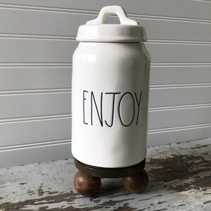 May include: White ceramic jar with a lid and handle, featuring the word "ENJOY" in black script. The jar sits on a dark wooden base with three rounded feet. The background is a white paneled wall.