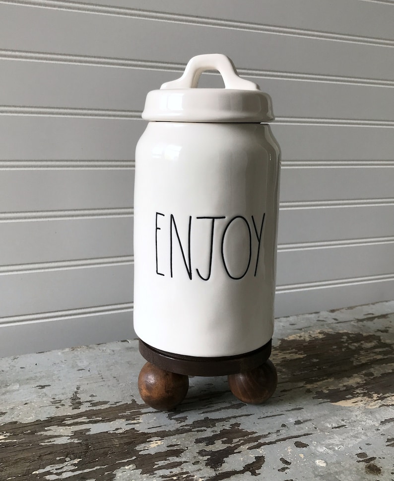 May include: White ceramic jar with a lid and handle, featuring the word "ENJOY" in black script. The jar sits on a dark wooden base with three rounded feet. The background is a white paneled wall.