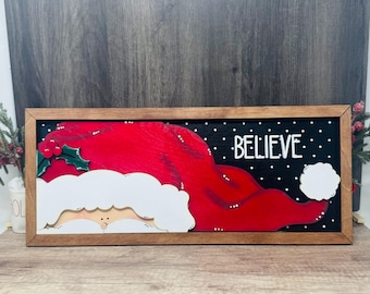 May include: Wooden framed Christmas sign featuring Santa's hat in red with white trim and a white beard. The word "BELIEVE" is written in white on a black background with white dots. The sign is a festive holiday decoration.