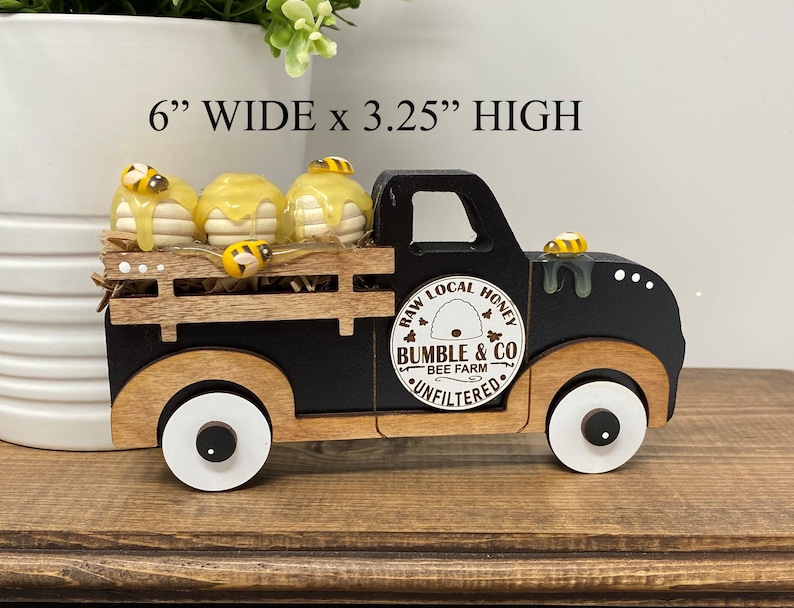 May include: A black and wood-toned decorative truck carrying three honey-covered treats and bee figurines. The truck is 15.24 cm wide and 8.26 cm high, with the text "RAW LOCAL HONEY" and "BUMBLE & CO BEE FARM UNFILTERED" on the side.