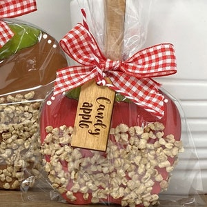 May include: A red candy apple with a wooden stick, coated in caramel and topped with nuts. The apple is wrapped in clear cellophane and tied with a red and white gingham bow. A wooden tag reads "Candy apple."