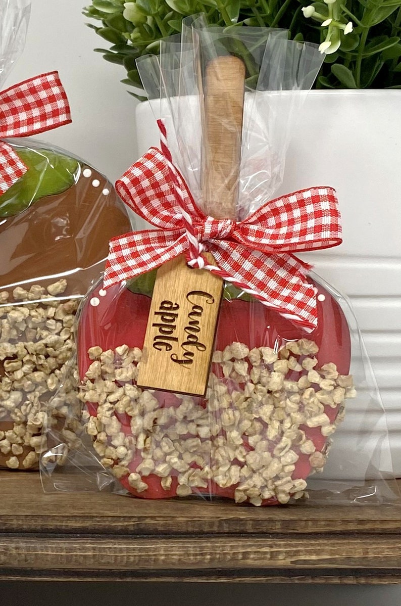 May include: A red candy apple with a wooden stick, coated in caramel and topped with nuts. The apple is wrapped in clear cellophane and tied with a red and white gingham bow. A wooden tag reads "Candy apple."