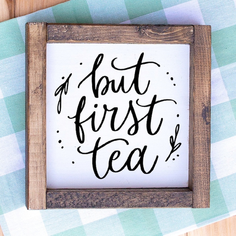May include: Square wooden sign with a white background and a dark brown frame. The sign features the phrase "but first tea" in elegant black script. The sign is placed on a green and white checkered cloth.