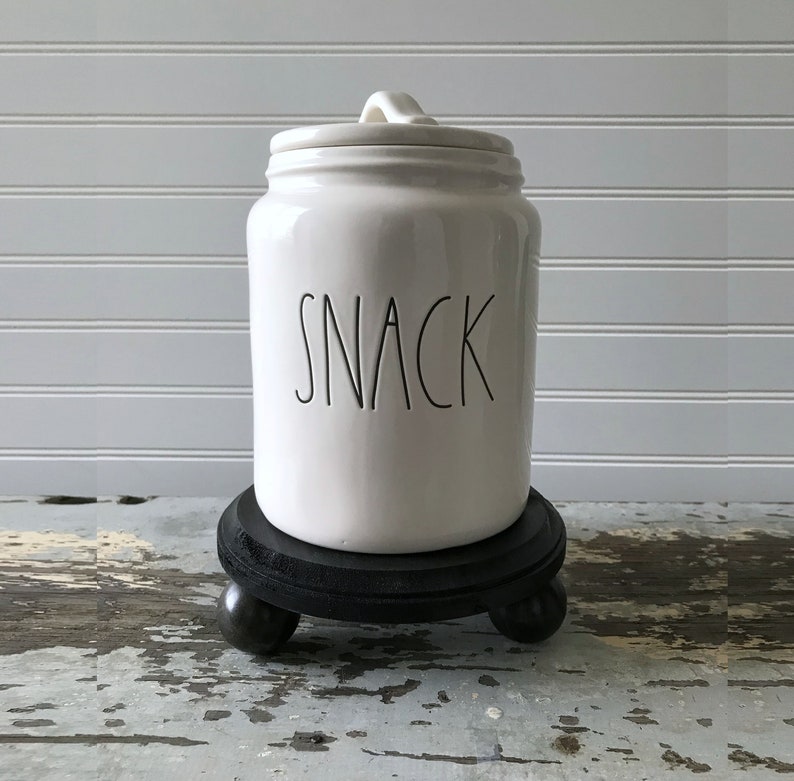 May include: A white ceramic snack jar with a lid and handle, featuring the word "SNACK" in black lettering. The jar sits on a small, round, black wooden stand with three ball feet. The background is a white, slatted wall.