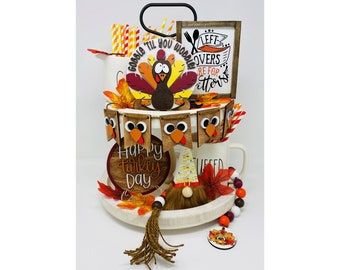 May include: A tiered tray decoration with a Thanksgiving theme. It features a turkey, autumn leaves, and a banner that says "Happy Turkey Day." The tray also includes a small wooden sign that says "Leftovers are for Eaters."