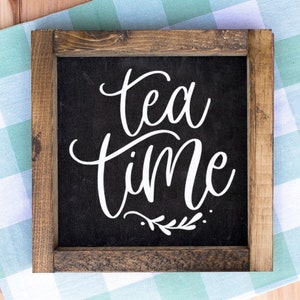 May include: Square wooden sign with a dark background and the words "tea time" in white script. The sign is framed with a rustic brown wood. A small sprig of leaves is below the word "time".