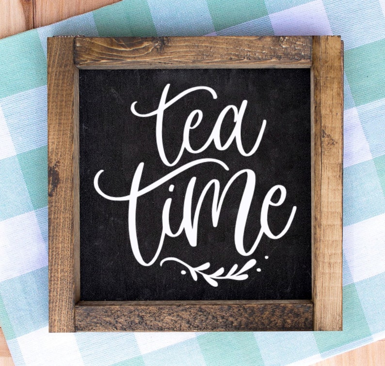 May include: Square wooden sign with a dark background and the words "tea time" in white script. The sign is framed with a rustic brown wood. A small sprig of leaves is below the word "time".