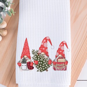 May include: White waffle-weave kitchen towel featuring three Christmas gnomes. Each gnome wears a red hat with snowflakes and holds festive items like a Christmas tree, mug, and a sign that says "Happy Holidays".