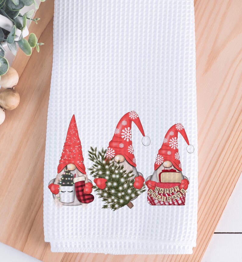 May include: White waffle-weave kitchen towel featuring three Christmas gnomes. Each gnome wears a red hat with snowflakes and holds festive items like a Christmas tree, mug, and a sign that says "Happy Holidays".