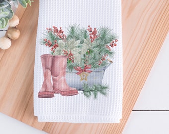 May include: White waffle-weave kitchen towel featuring a watercolor design. The design includes pink rain boots, a galvanized metal bucket filled with greenery, red berries, and a bow. A gold star-shaped ornament hangs from the bucket.