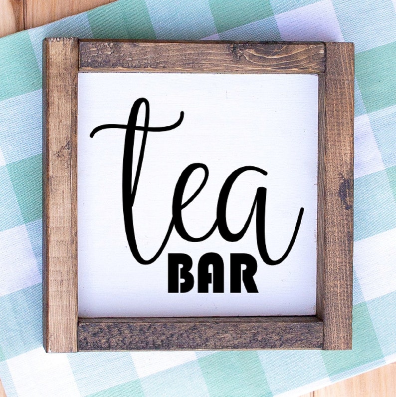May include: Square wooden sign with a white background and the words "Tea Bar" in black script and block letters. The sign is framed with dark brown wood and rests on a green and white checkered cloth.
