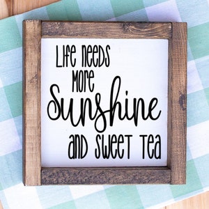 May include: Square wooden sign with a white background and a dark brown frame. The sign features the words "Life needs more sunshine and sweet tea" in black script and block letters. The sign is placed on a light blue and white checkered surface.