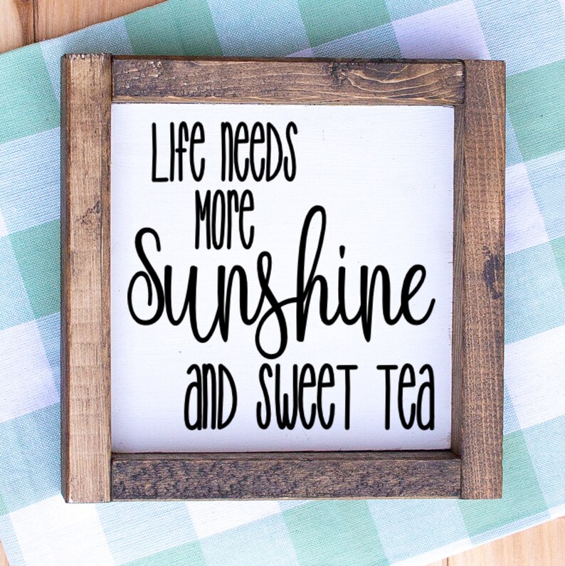 May include: Square wooden sign with a white background and a dark brown frame. The sign features the words "Life needs more sunshine and sweet tea" in black script and block letters. The sign is placed on a light blue and white checkered surface.