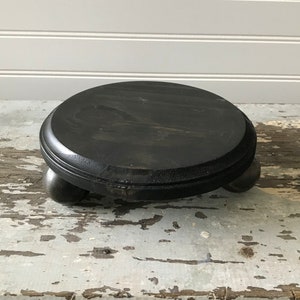 May include: A round, black wooden pedestal with a dark finish. The pedestal has a flat, circular top and is supported by three small, rounded feet. The wood grain is visible, adding texture to the piece. This decorative stand is suitable for displaying small items.
