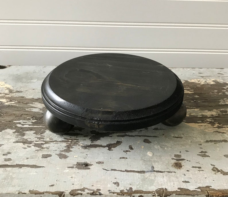 May include: A round, black wooden pedestal with a dark finish. The pedestal has a flat, circular top and is supported by three small, rounded feet. The wood grain is visible, adding texture to the piece. This decorative stand is suitable for displaying small items.