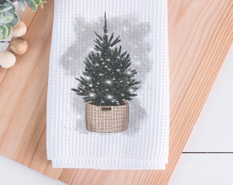 May include: White waffle-weave kitchen towel featuring a Christmas tree illustration. The tree is dark green with twinkling lights, set in a woven basket. The design has a watercolor effect, perfect for holiday decor.