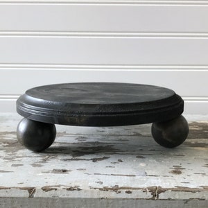 May include: A small, round, black wooden plant stand with a circular top and three spherical feet. The stand is painted black and has a distressed finish, giving it a rustic appearance. The stand is suitable for displaying small plants or decorative items.