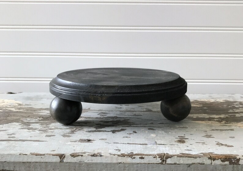 May include: A small, round, black wooden plant stand with a circular top and three spherical feet. The stand is painted black and has a distressed finish, giving it a rustic appearance. The stand is suitable for displaying small plants or decorative items.