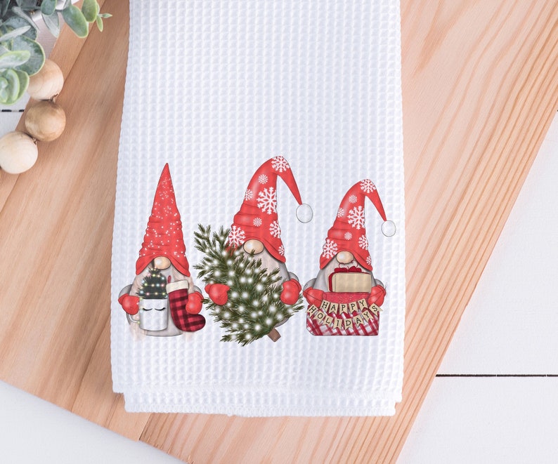 May include: White waffle-weave kitchen towel featuring three gnomes in red hats with white pom-poms. The gnomes are holding a Christmas tree, a mug, and a sign that says "Happy Holidays".