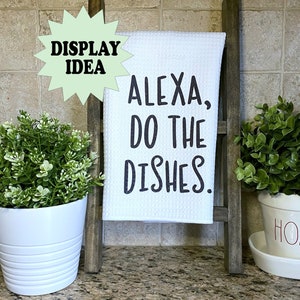 May include: A white kitchen towel with the text "ALEXA, DO THE DISHES" in black lettering is displayed on a small wooden ladder. The ladder is gray and stands on a countertop. Potted plants flank the ladder.