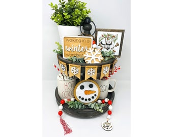 May include: A tiered tray with winter-themed decorations. Includes a small sign that says "walking in a winter wonderland", a snowman, snowflake, and a banner with snowflakes. There are two mugs that say "Hot Cocoa" and a string of beads.