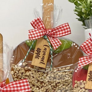 May include: Caramel apple on a wooden stick, wrapped in clear cellophane. The caramel apple is covered in nuts and has a red and white gingham bow. A wooden tag reads "Caramel apple".