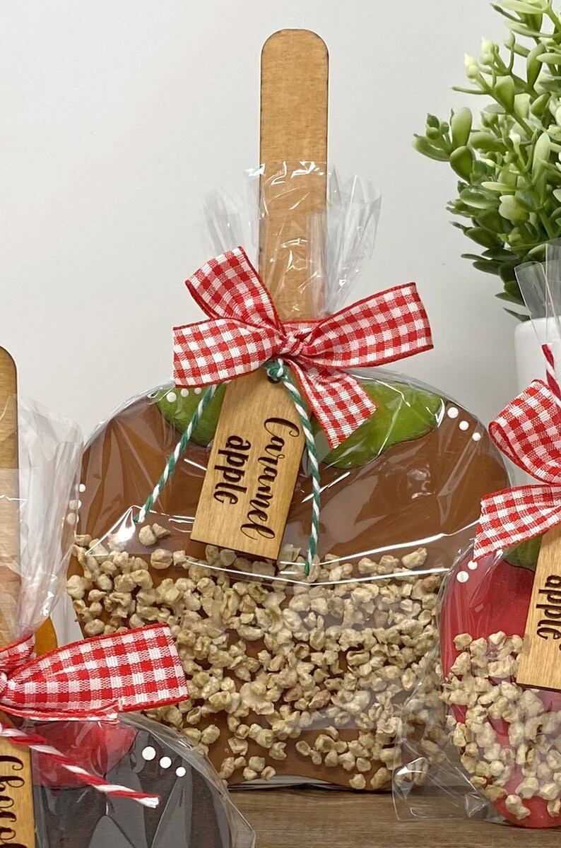 May include: Caramel apple on a wooden stick, wrapped in clear cellophane. The caramel apple is covered in nuts and has a red and white gingham bow. A wooden tag reads "Caramel apple".