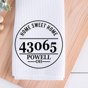 May include: White waffle-weave kitchen towel with a black circular design that reads "HOME SWEET HOME" above the numbers "43065" and "POWELL OH" below. The towel is on a wooden cutting board.
