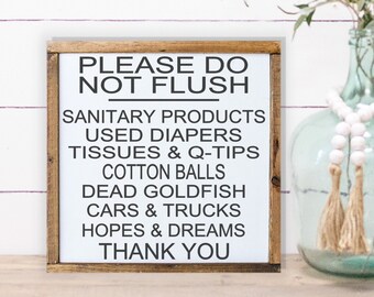 May include: A framed wooden sign with white background and black text that reads "PLEASE DO NOT FLUSH" followed by a list of items. The sign is on a wooden surface with a blurred background.