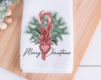 May include: White waffle-weave Christmas hand towel with a festive design. The design features a candy cane, red berries, green pine needles, and a red heart-shaped ornament. The words "Merry Christmas" are written in black script.