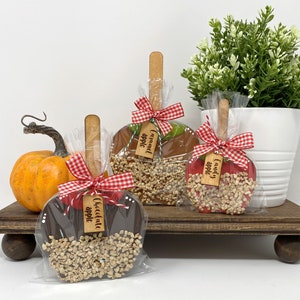 May include: Three caramel apples on a wooden tray, each with a wooden stick and a red and white checkered bow. The apples are labeled "Chocolate apple", "Caramel apple", and "Candy apple". The apples are covered in nuts and wrapped in clear cellophane.
