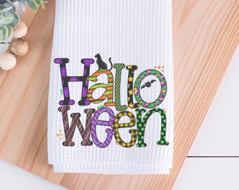 May include: White waffle-weave kitchen towel with the word "Halloween" in colorful, patterned letters. A black cat sits atop the "H", and a bat graphic is in the "O". The towel is displayed on a wooden surface.