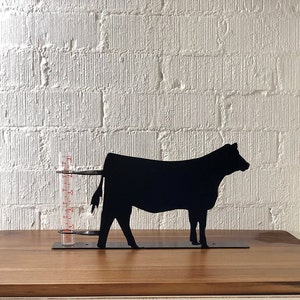 May include: A black metal cow silhouette rain gauge with a clear, cylindrical measuring tube. The gauge has red measurement markings and sits on a rectangular base. The background is a white brick wall.