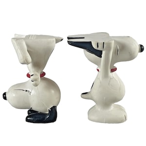 May include: Two Snoopy figurines. Each figurine is white with black accents on the ears, feet, and nose. They have a red collar. One figurine is facing left, and the other is facing right. The figurines are on a white background.