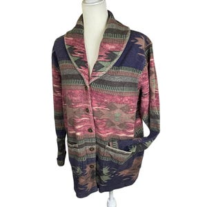 May include: A long-sleeved button-down jacket with a shawl collar. The jacket features a Southwestern-inspired pattern in shades of pink, blue, green, and brown. It has two front pockets and is displayed on a mannequin.
