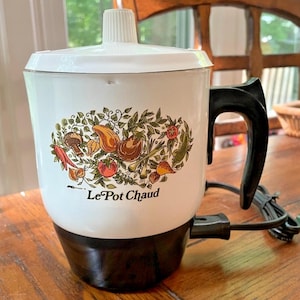 May include: A white electric pot with a black base and handle, featuring a white lid with a central knob. The front displays a colorful illustration of various vegetables and the text Le Pot Chaud in black lettering, with a black power cord attached.