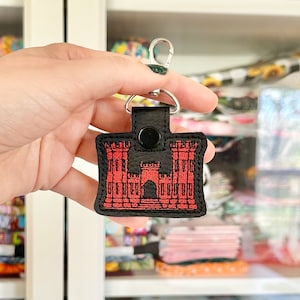 May include: A black leather keychain with a red embroidered image of a castle. The keychain has a silver metal clasp.