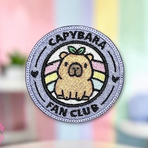 May include: A circular embroidered patch featuring a tan capybara with a small green leaf on its head. The capybara is centered in front of a pastel rainbow. The border is light purple with the black text CAPYBARA FAN CLUB and two small hearts.