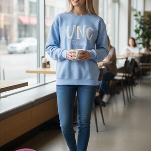 Op de afbeelding: Een lichtblauwe sweatshirt met witte letters "UNC Tarheels". De persoon draagt een blauwe spijkerbroek en witte sneakers, en houdt een witte mok vast. De foto is binnenshuis genomen, met een raam en tafels op de achtergrond.