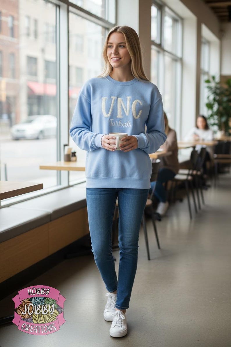 Op de afbeelding: Een lichtblauwe sweatshirt met witte letters "UNC Tarheels". De persoon draagt een blauwe spijkerbroek en witte sneakers, en houdt een witte mok vast. De foto is binnenshuis genomen, met een raam en tafels op de achtergrond.