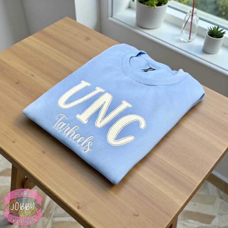 Op de afbeelding: Een lichtblauwe sweatshirt met de letters "UNC" en het woord "Tarheels" in wit geborduurd. De sweatshirt is netjes op een houten tafel gevouwen. Het ontwerp is gecentreerd op de borst.