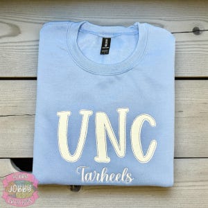 Op de afbeelding: Een lichtblauw sweatshirt met de witte, gestikte letters "UNC". Daaronder is het woord "Tarheels" in het wit geborduurd. Het sweatshirt is netjes op een houten oppervlak gevouwen.