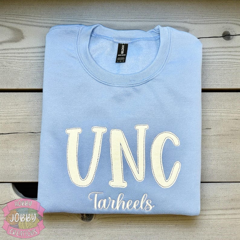 Op de afbeelding: Een lichtblauw sweatshirt met de witte, gestikte letters "UNC". Daaronder is het woord "Tarheels" in het wit geborduurd. Het sweatshirt is netjes op een houten oppervlak gevouwen.