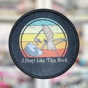 May include: Round patch with a black border featuring a cartoon creature and a rock against a sunset background. The text "I Don't Like This Rock" is printed below the image. The patch is a fun accessory.