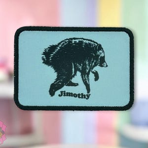 May include: A rectangular patch with a light blue background and a black border. The patch features a black graphic illustration of a bear walking forward, with the name Jimothy printed in black text underneath the bear's hind legs.